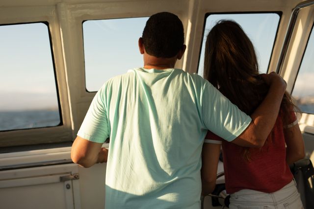 Rear view of Caucasian man and his teenage daughter enjoying time together on holiday in the sun by the coast, standing in a boat, embracing admiring the view.