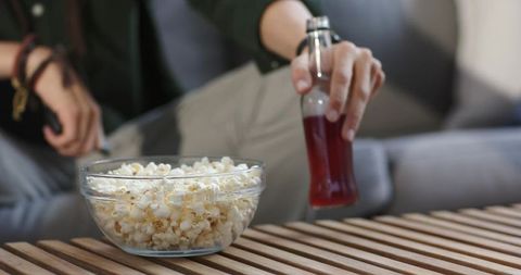 Mid-adult man relaxing on couch holding remote and bottle beside bowl of popcorn