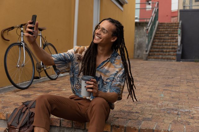 Biracial smiling alternative man with dreadlocks out and about in the city on a sunny day, using his smartphone taking selfies, bike next to him. Urban trendy man on the go.