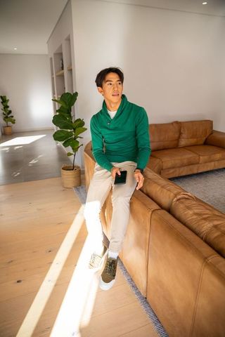 Young asian man wearing green sweater sitting on tan leather sofa with smartphone in modern home
