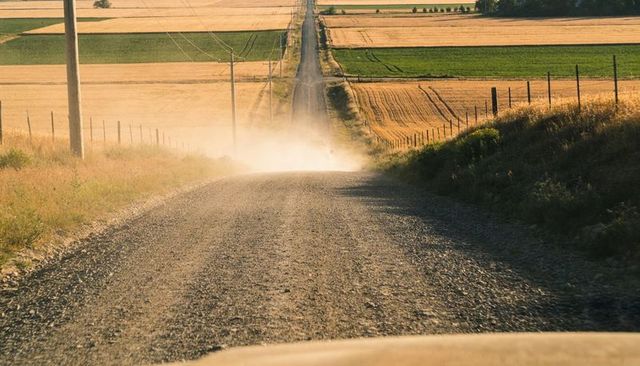 Endless gravel road running through golden farmland at late light leaving dust trail