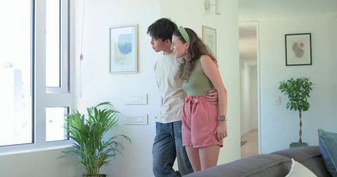 Young couple admiring sunlit window in modern minimalist living room with plants and smartwatch