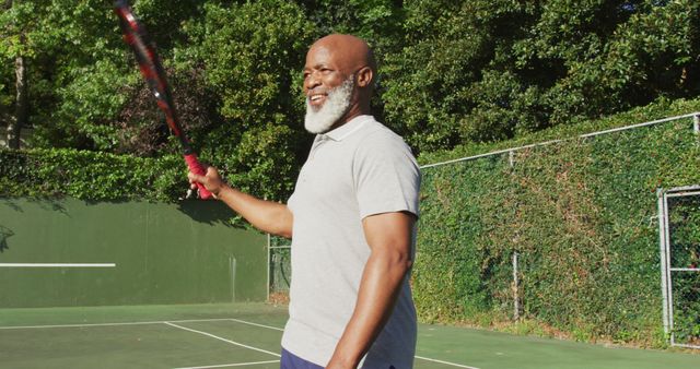 A senior man is standing on an outdoor tennis court, holding a tennis racket and smiling. He appears happy and engaged in the sport. This could be used to illustrate senior fitness, active lifestyles, or the enjoyment of recreational sports. The background shows trees and a fence, indicating a peaceful setting for outdoor activities.