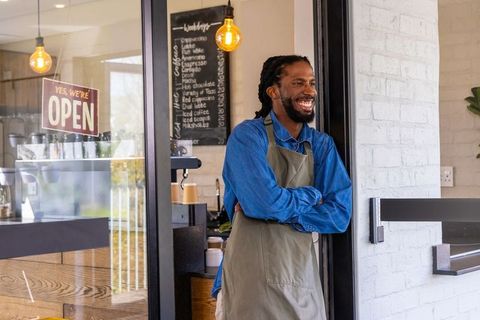 Smiling african american barista leaning in cafe doorway with open sign and cozy interior