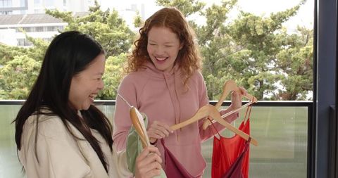 Friends comparing dresses on balcony, smiling holding wooden hangers, casual urban fashion
