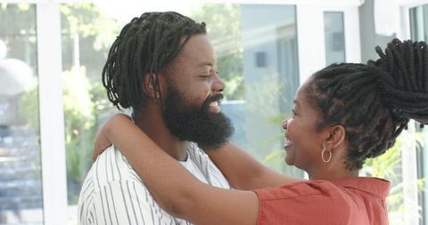 African american couple embracing, gazing and smiling by sunlit glass doors at home
