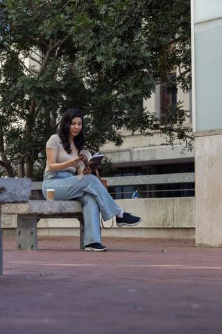 Woman reading paperback on urban bench with coffee and shoulder bag in leafy city plaza