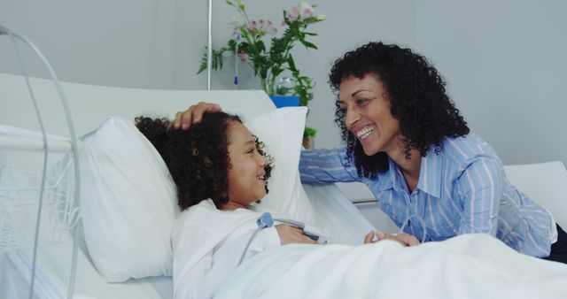 Mother lovingly interacting with her child who is lying in a hospital bed, both smiling warmly. Ideal for showcasing family support during healthcare situations, patient care and recovery, emotional bonds, and the importance of familial support in medical settings.