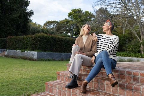 Mature friends laughing while sitting on red brick steps in garden wearing ankle boots