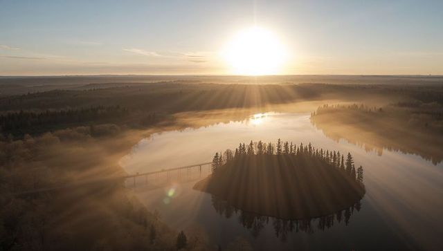 Golden sunrise rays over misty lake islet with bridge and forest reflection, aerial serene