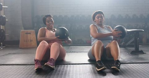 Backlit black women doing medicine ball russian twists together in gym workout