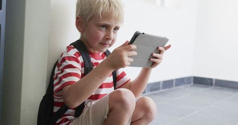 Blond boy sitting on hallway floor using tablet with backpack and focused expression