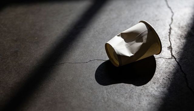 Crushed paper cup lying on cracked concrete, casting oval shadow in diagonal light