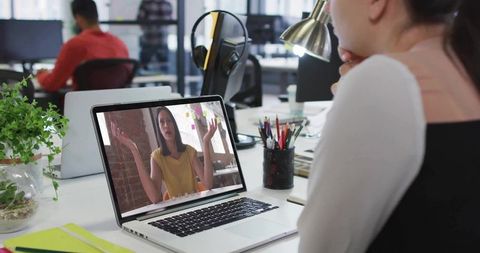 Woman attending virtual meeting on laptop at office desk with plant and headphones