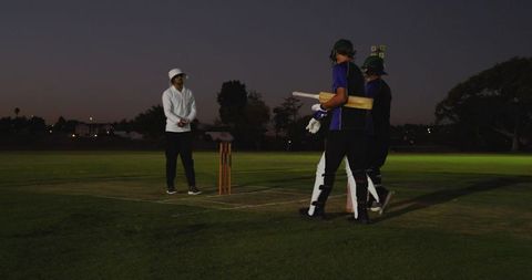 Twilight cricket scene two batsmen walking past stumps with umpire watching