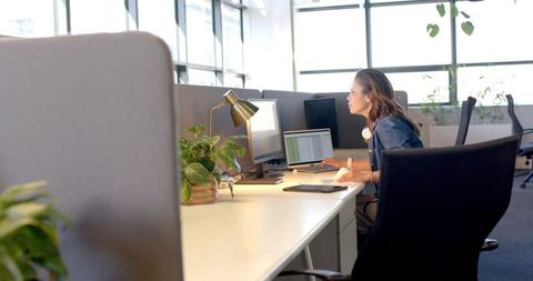 Mid-adult woman working on laptop in bright modern office with plants and dual monitors