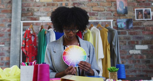 Black woman holding a color palette, surrounded by vibrant fabric rolls and clothing on hangers. Background features a brick wall with artistic photos. Ideal for topics on fashion design, creative professions, color theory, and artistic inspiration.