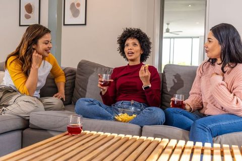 Three friends chatting and snacking on sofa, casual home gathering with drinks, laughter