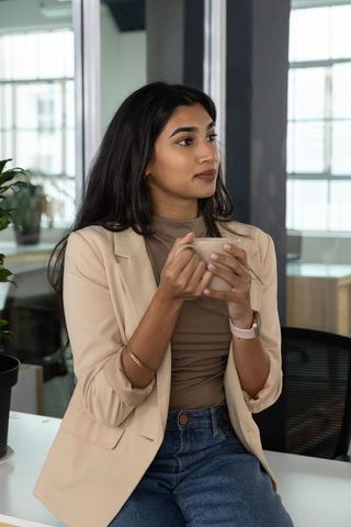 Indian businesswoman sipping coffee on office counter while looking thoughtful in coworking