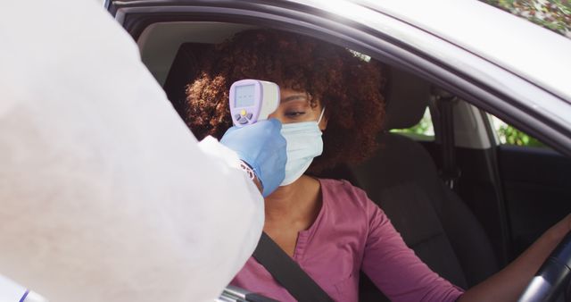 Healthcare worker using infrared thermometer to check temperature of woman sitting in car. Woman wearing face mask, ensuring safety and health during pandemic. Suitable for articles on drive-thru testing, COVID-19 safety measures, and healthcare services.