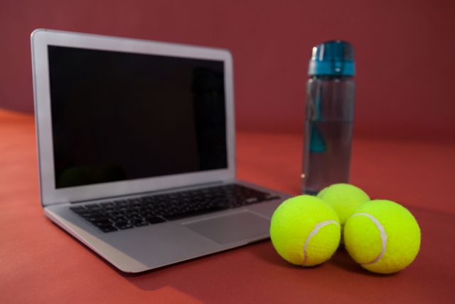 Close up of tennis balls by laptop and water bottle against maroon background