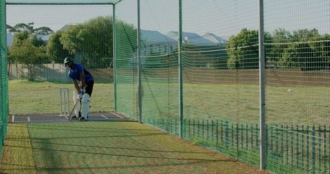 Indian cricketer practicing in net enclosure on sunny field wearing pads and helmet