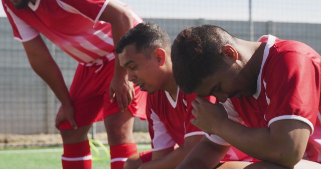 Three football players wearing red uniforms are sitting on the field, reflecting on a disappointing match. They appear downcast, possibly after losing a game. Their defeated posture and facial expressions highlight their emotional response to the outcome. This image can be used to depict themes of defeat, sportsmanship, team dynamics, and determination.