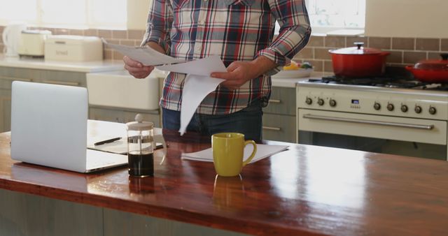 A middle-aged Caucasian man stands in a kitchen, reviewing papers with a laptop and a cup of coffee on the counter, with copy space. His casual attire and the domestic setting suggest he may be working from home or managing household finances.