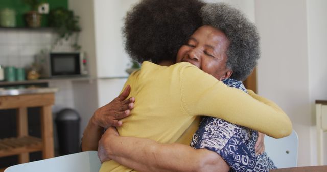 Happy african american adult daughter and senior mother hugging in kitchen at home, slow motion. Family, relaxation, togetherness, love, motherhood and domestic life, unaltered.