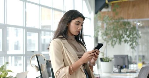 Indian businesswoman checking smartphone in modern bright office with laptop and indoor plants