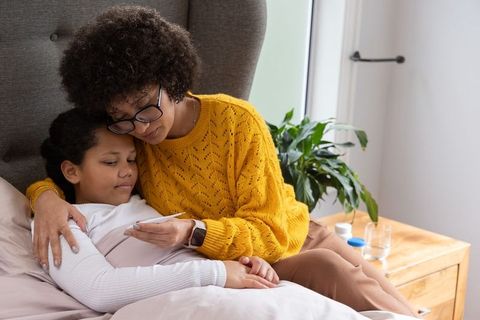 African american mother comforting sick daughter while checking temperature in bedroom