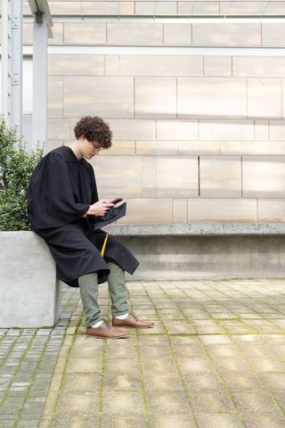 Teen graduate sitting on modern campus bench checking smartphone in black gown with tassel