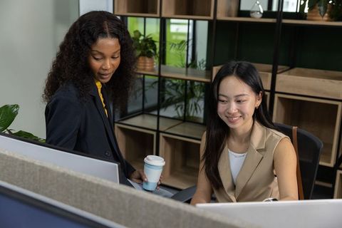 Female coworkers collaborating at office desk, using monitors and holding coffee