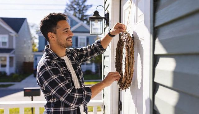 Hispanic man hanging grapevine wreath on sunny suburban porch for seasonal decor