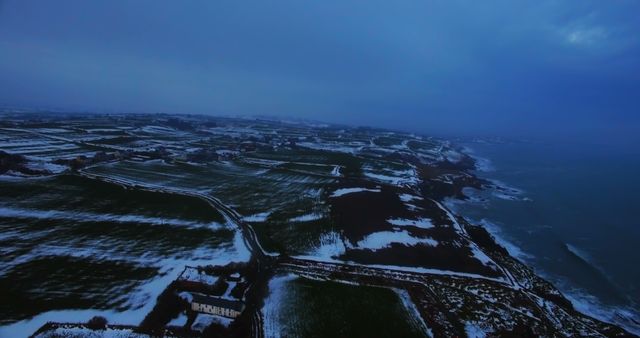 Aerial view captures expansive snow-covered farmland meeting the coastline during dusk. Deep blue hues and falling darkness enhance the serene, almost mystical atmosphere of this winter landscape. Ideal for use in travel blogs, winter seasonal promotions, agricultural marketing materials, and scenic nature collections.