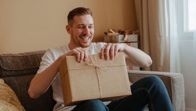 Smiling man holding wrapped kraft parcel on sofa, unwrapping gift at home on sunny day