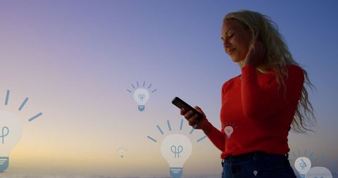 Creative woman using smartphone with digital bulb icons at sunset