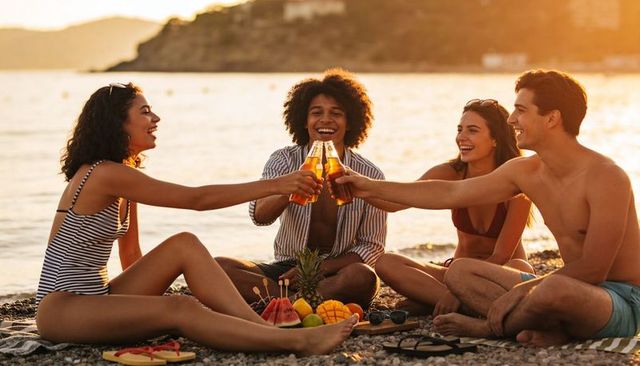 Friends toasting bottles at sunset on pebble beach with pineapple, mango and watermelon