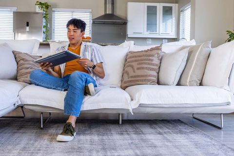Asian man relaxing on white sofa using tablet in modern open-plan living room