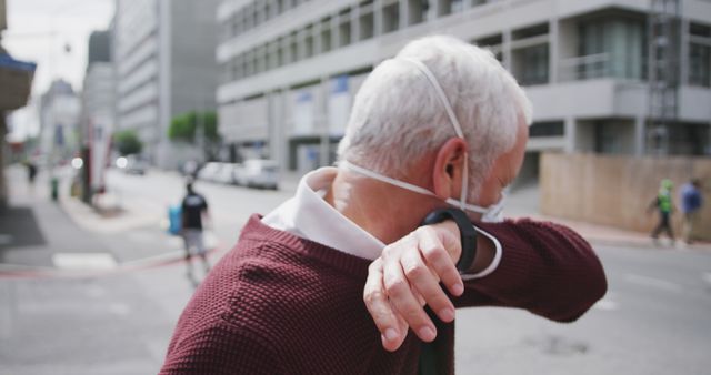 Elderly man coughs into elbow while wearing face mask in city street, emphasizing health precautions and safety measures. Ideal for content on public health, pandemic response, face mask usage and respiratory health-related awareness.
