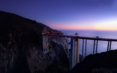 Twilight coastal arch bridge glowing with car light trails over Pacific coastline