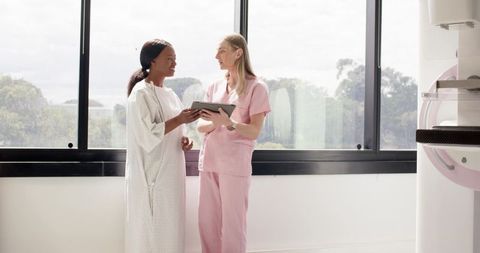 Radiology technician consulting Black woman patient in gown with tablet near mammography machine