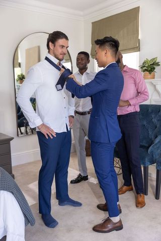 Diverse groomsmen helping groom tie necktie in bright bedroom before wedding
