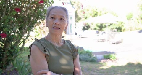 Senior woman standing in sunlit backyard garden looking thoughtful with arms crossed