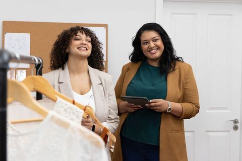 Diverse female designers reviewing lace garments on rack while using tablet for planning