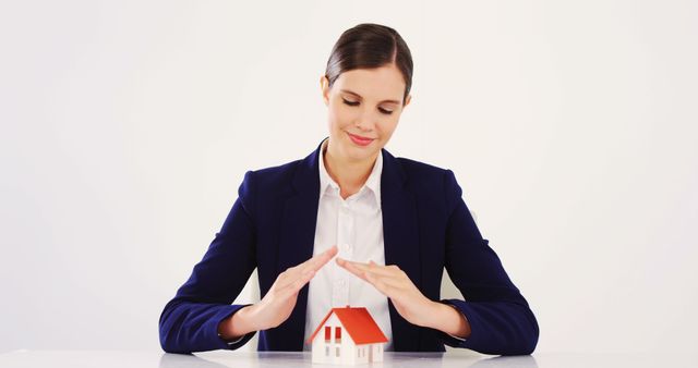 A young Caucasian woman in professional attire is protectively placing her hands around a small model house, with copy space. Her gesture suggests a concept of home insurance or real estate protection.