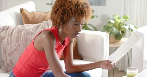 African american woman reading invoice on bright living room sofa wearing red sleeveless top and jea