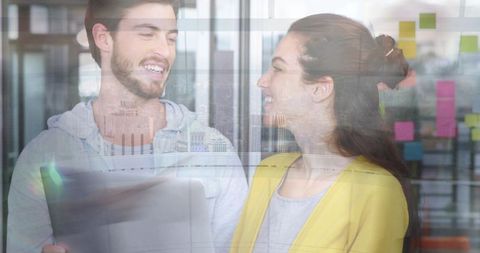 Smiling young colleagues collaborating with laptop by glass wall with colorful sticky notes
