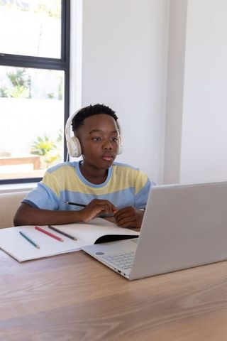African american boy studying online wearing headphones at home desk with laptop