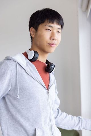 Thoughtful asian young man wearing headphones standing by window in gray hoodie